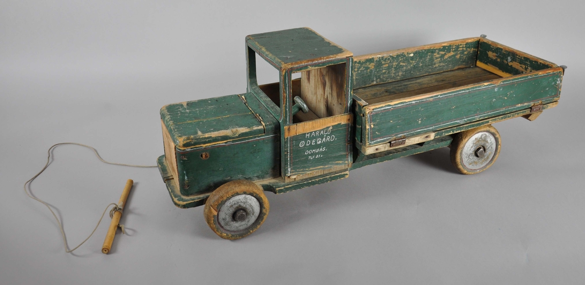 Håndlaget lastebil av tre. Består av frampart og lasteplan, samt bevegelige hjul. Lastebilen har en grønn grunnfarge med røde dekorstriper. Malingen er avslitt flere steder. Lasteplan og panser er hengslet slik at det kan tippes. Bak- og sidedør på lasteplanet er også hengslet slik at de kan legges ut. På framsiden står det en pinne ut på høyre side, på venstre side skal det være en tilsvarende pinne, men denne har knekt. På framsiden er det festet en tråd med pinne i enden, for å dra lastebilen.