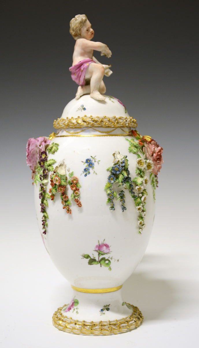Potpourrikrukke av porselen med stett og lokk. Lokket er dekorert med håndmalt blomsterdekor og langs kanten en bord med blader i tre høyder dekorert med gull, over bladene gullfargede små kuler. På toppen av lokket en kjerub eller kupid som sitter på sitt ene kne. Han har på seg et rosa lendeklede og har holdt i et skjold eller en atributt. 
Urnens korpus er dekorert med håndmalte blomster, og modellerte blomster som sitter i klynger og strekker seg nedover korpus. De modellerte blomstene er uglasert og håndmalt. Urnen mangler en hanke. Den eksisterende hanken har modellerte blomster ved festene som strekker seg nedover i klynger.  Langs stettens kant samme bladbord som på lokket.