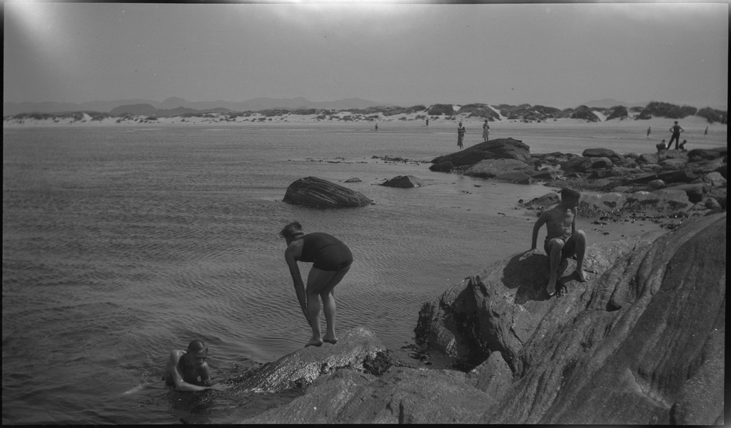 En stor vennegjeng i lystig lag på stranda på Sola. De har trommer og trekkspill og leker i vannet og på land med og uten ball. Sola strandhotell ligger i bakgrunnen. Tor Johannessen og Harald Bergsaker er på bilde nr. 14 og 18.
