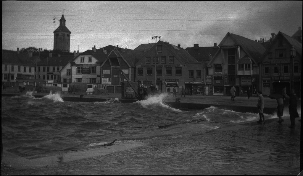 Bølger slår inn over Skagenkaien i Stavanger. En mann prøver å fiske opp noe med en lang stang og en mann vinsjer opp en stor stålbjelke, mens flere ser på.