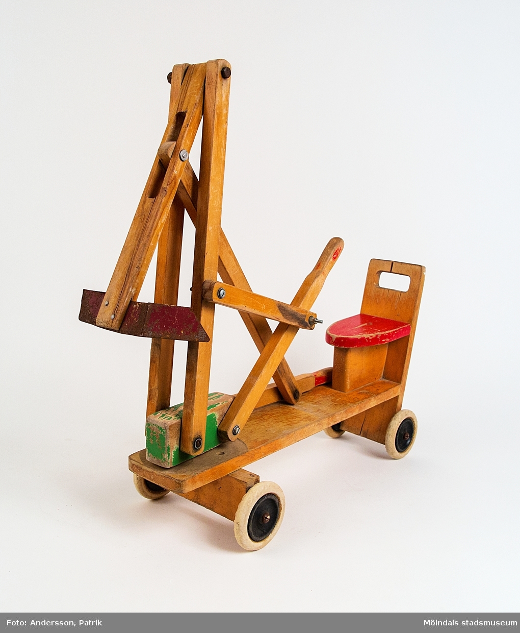 Grävskopa. Tillverkad av klarlackat trä med färglagda detaljer och röd sits. Fyra hjul med gummidäck. Grävskopan regleras av ett barn med hjälp av två spakar för lek i sandlåda.