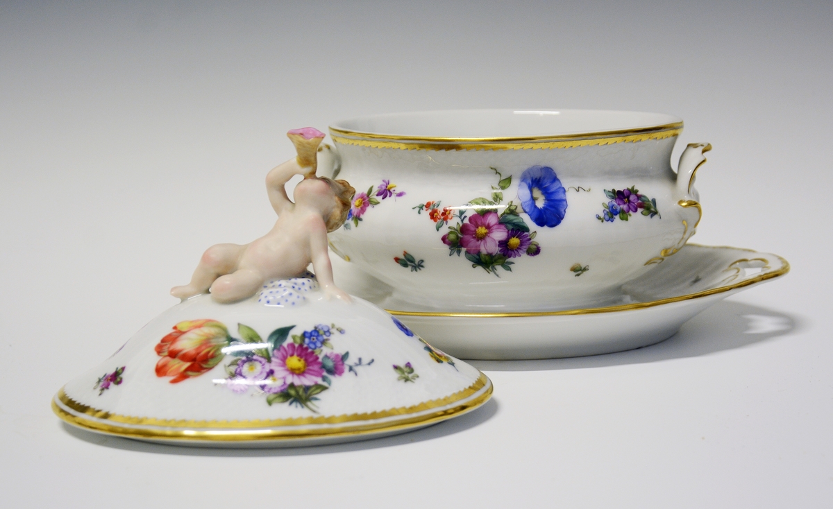 Sauseterrin av porselen dekrorert med håndmalt blomsterdekor og modellert figur av et barn med blåseintstrument som lokknapp. Hanker på hver siden med skjellform. Alle kanter er markert med gull.