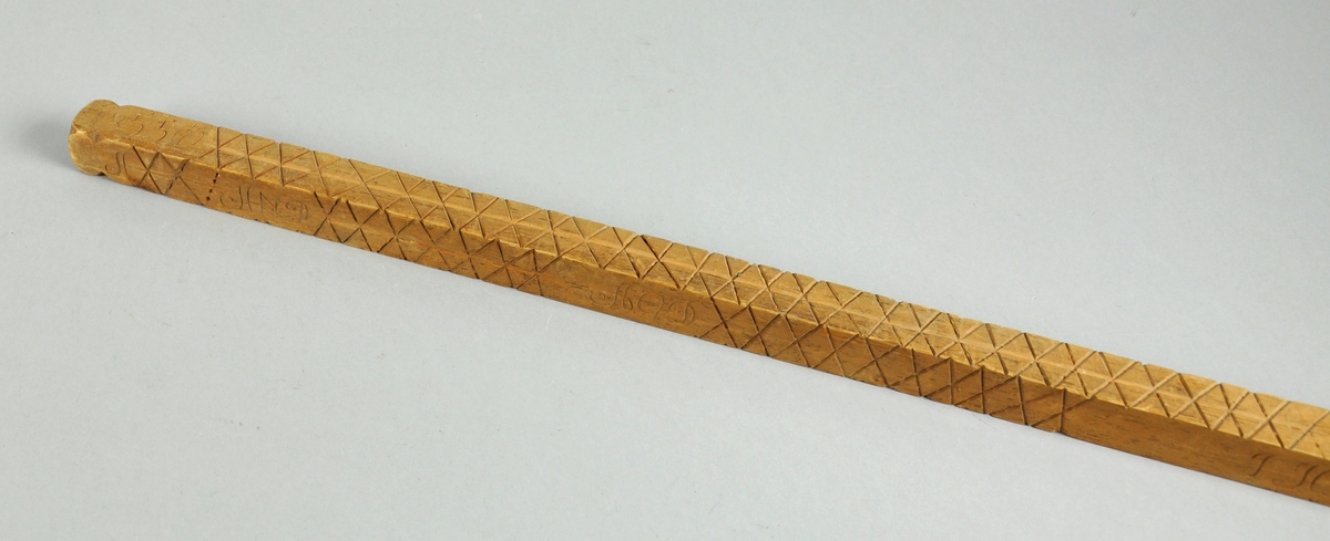 Målestokk av tre som er spiss i den ene enden. Det er inngravert mange kryss og punkter alle fire langsider med signaturer for hver rekke av merker.