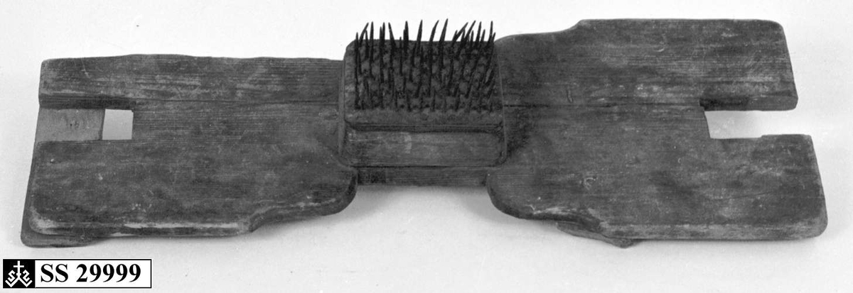 Linhekle av tre som er smalere på midten, der det er festet en trekloss med stålspiker. Trebrettet er skadet og reparert ved å spikre på to trestykker på tvers på undersiden.