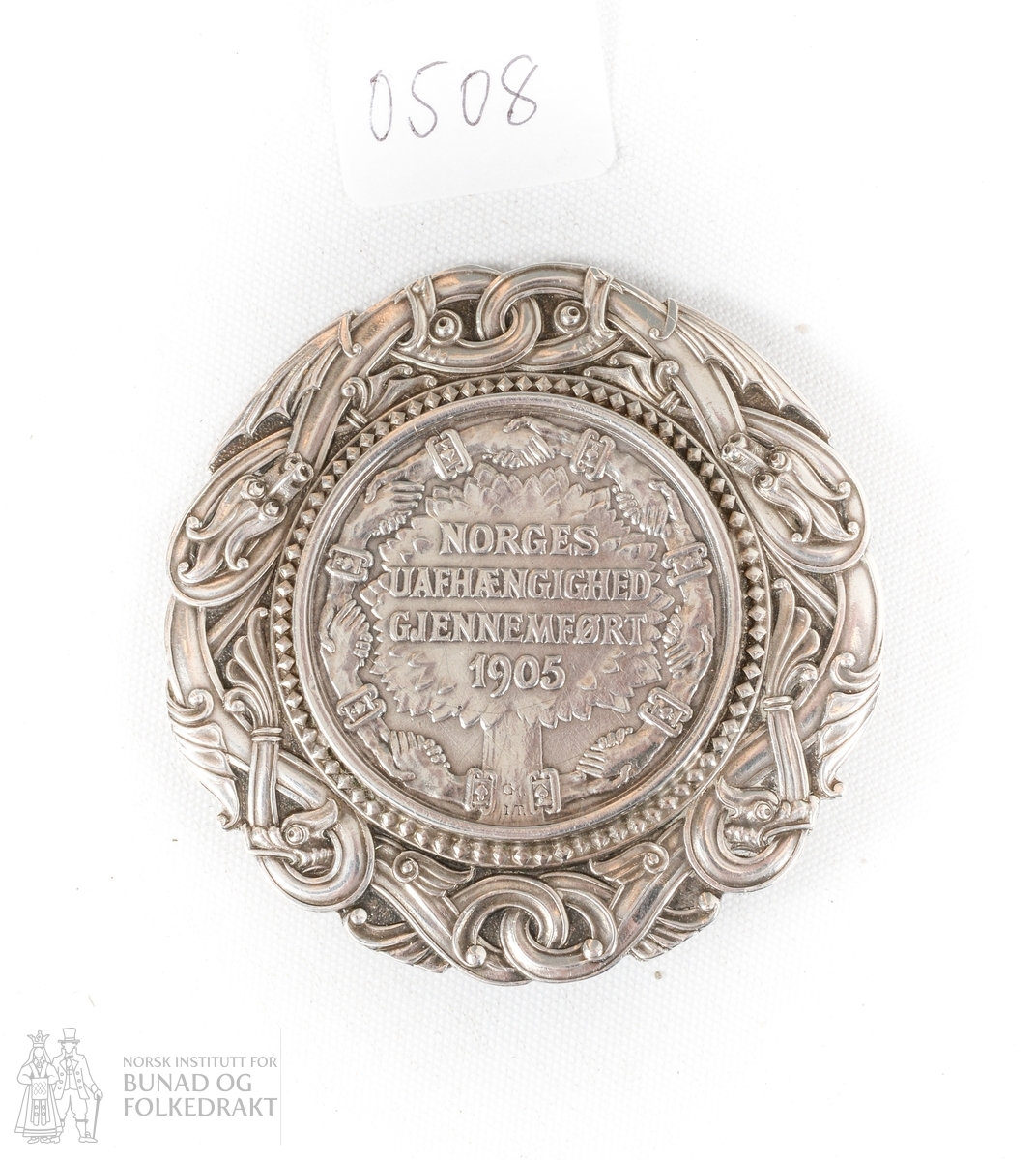 Nål med innfelt mynt. Dragemotiv rundt kanten av mynten. PÅ eine sida av mynten (baksida av nåla). 2 KRONER 19 x 06
På framsida av nåla: NORGES UAFHÆNGIGHED GJENNEMFØRT 1905