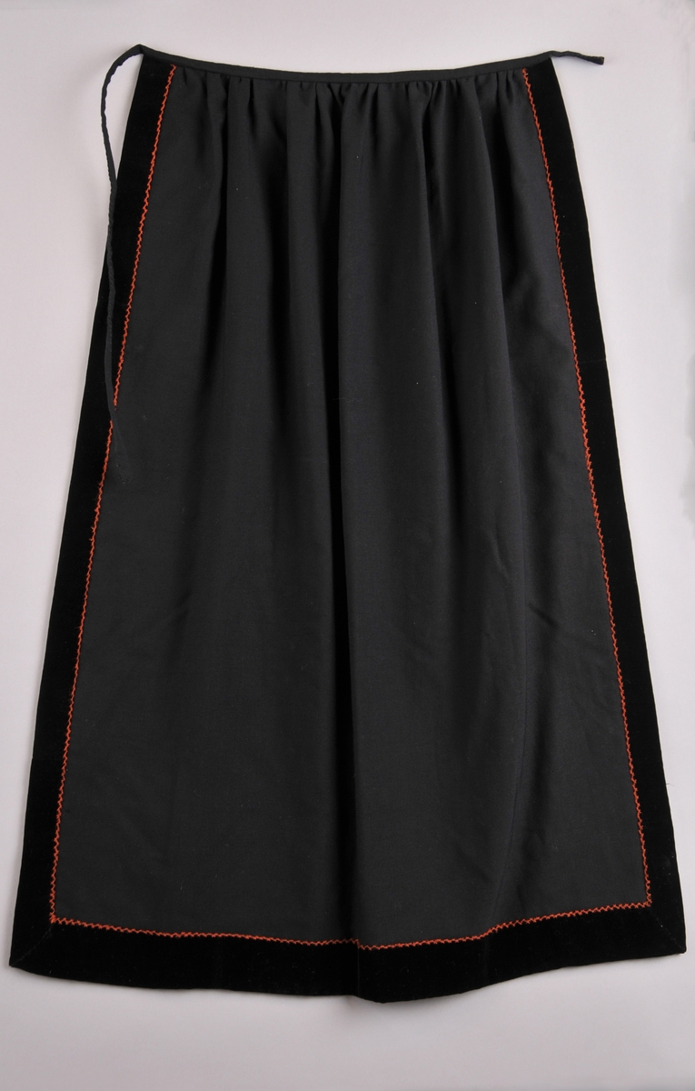 Forkle i svart ullsateng med svarte fløyelskantar rundt, og ein saum med raud-orange heksesting innanfor kanten.