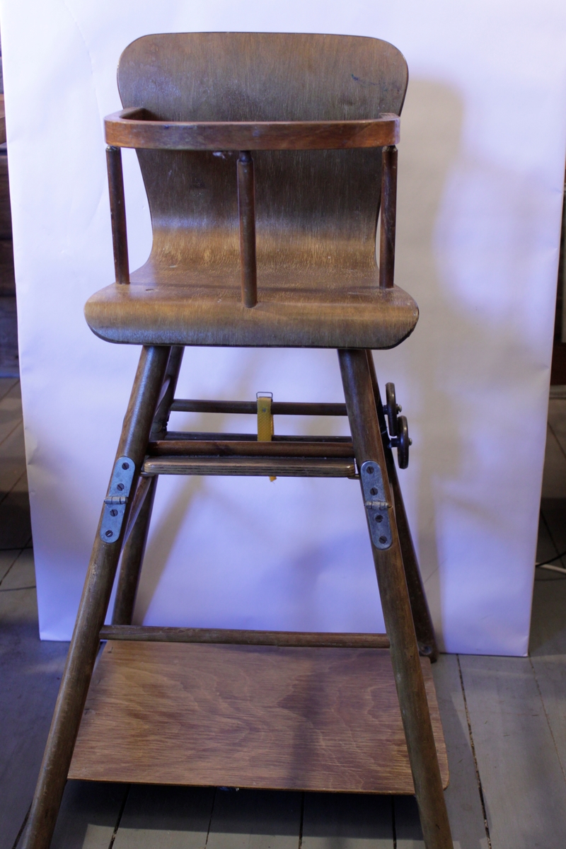 Hög barnstol med sits i böjträ. I mitten på de höga benen sitter gångjärn och små hjul, i botten en skiva. När man fäller benen vid gångjärnen får barnet ett bord på hjul till stolen.