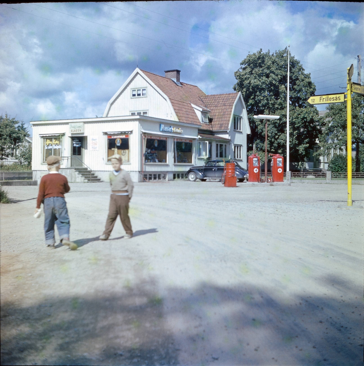 Två pojkar på gårdsplan framför Edvin Johanssons affär på Viskastigen 29, med cykel och "Fantomknarren" (Monark Blue Arrow M550) i skyltfönstren. Dan Johansson står med ryggen åt kameran och Bosse Eierborg till höger. Över dörren på gaveln sitter en skylt för Dalstrands kläder. Vid butiken finns två röda bensinpumpar med Caltexstjärnan och en parkerad Opel Admiral 1939.