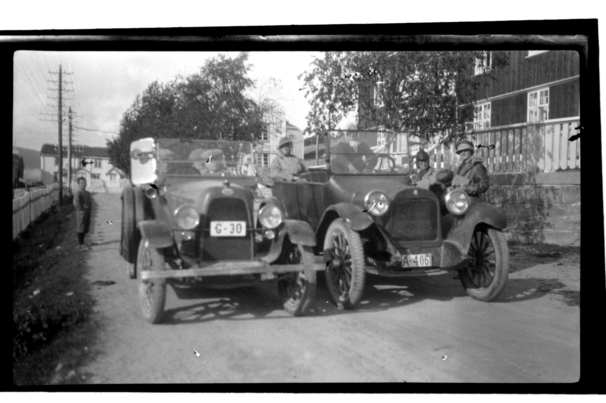 To biler, eid av henholdsvis Halfdan og Rolf Sundt Sr., med passasjerer under rast på ukjent sted med store trebygninger. Fotografert 1925.