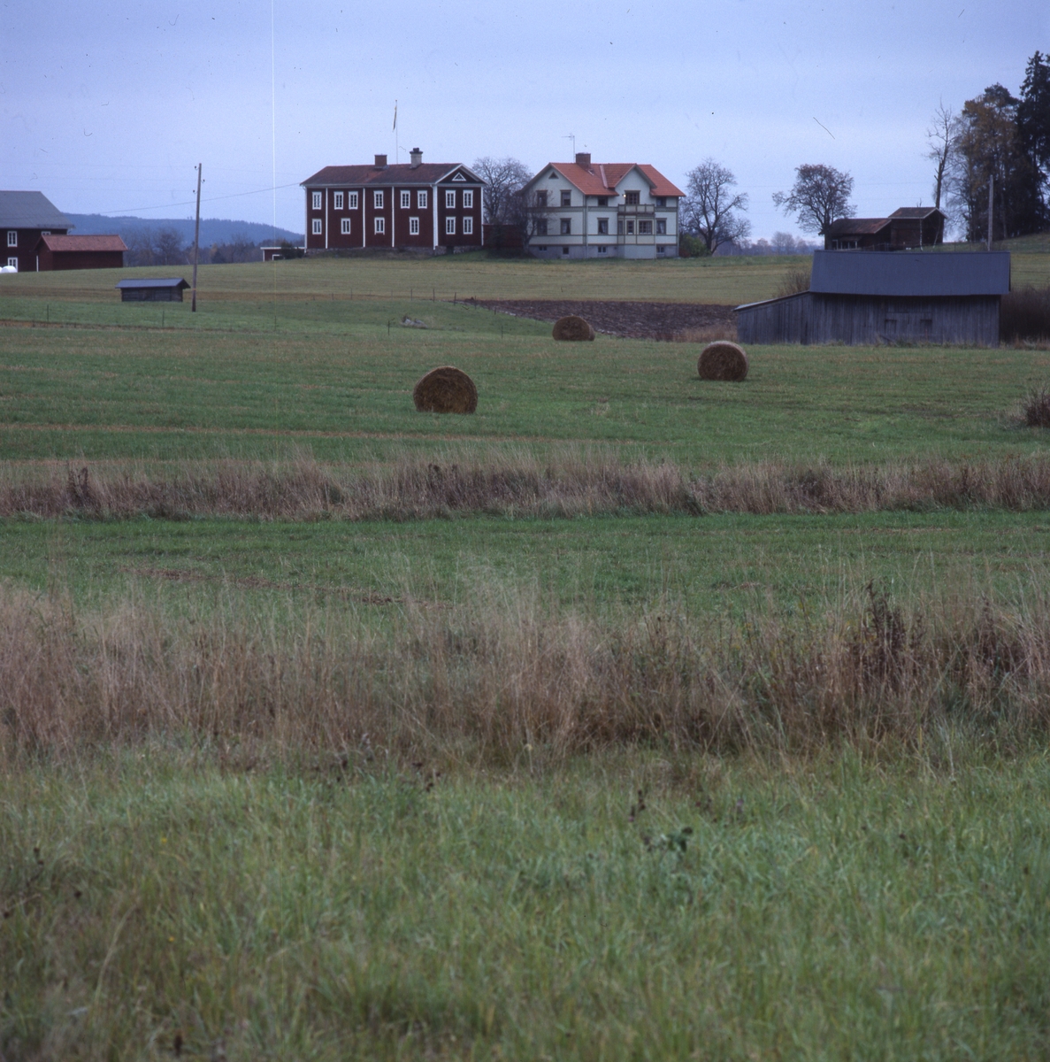 Halmbalar utspridda bland lador i ett odlingslandskap, med en gård med två boningshus på en höjd i bakgrunden, oktober 2001.