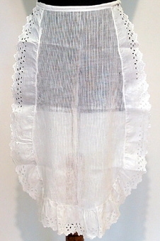 Serveringsförkläde av vitt smalrandigt bomullstyg. Runtom förklädet finns fastsydd en rynkad volang av bomullsbrodyr.