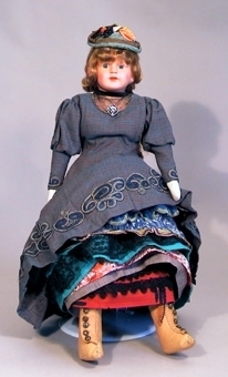 Docka med celluloidhuvud sydd omkring 1970 med kläder i 1890-talets stil. Blå-brun klänning och sex underkjolar, mamelucker samt kängor i ljust skinn.