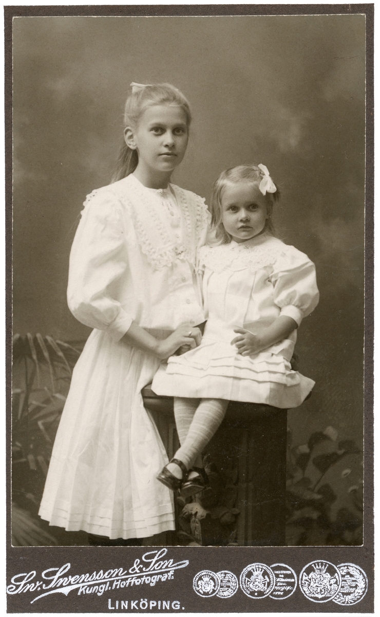 Porträtt av systrarna Hildur och Karin Eriksson. Syskonen var döttrar till makarna Erik och Lovisa Eriksson. Paret hade inflyttat till Linköping från Eskilstuna 1893. Familjen var bosatt i Tannefors där föräldrarna var kvarnägare och fadern etablerat rörelsen AB Erikssons Valskvarn.