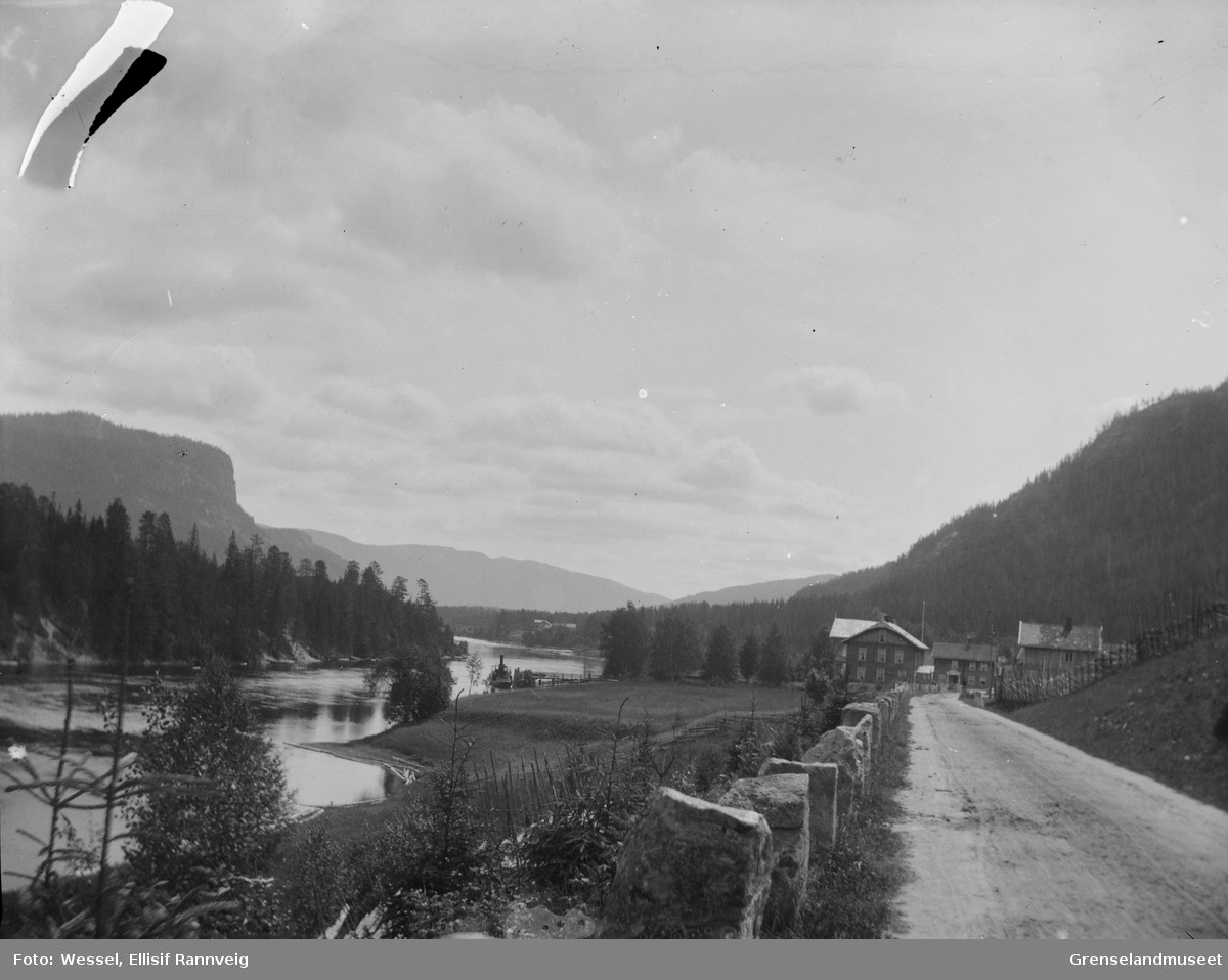 En vei frem mot en gårdsplass, store bygninger. En elv til venstre, en dampbåt på elven. Granskog. Trolig østlandet, ca 1901.