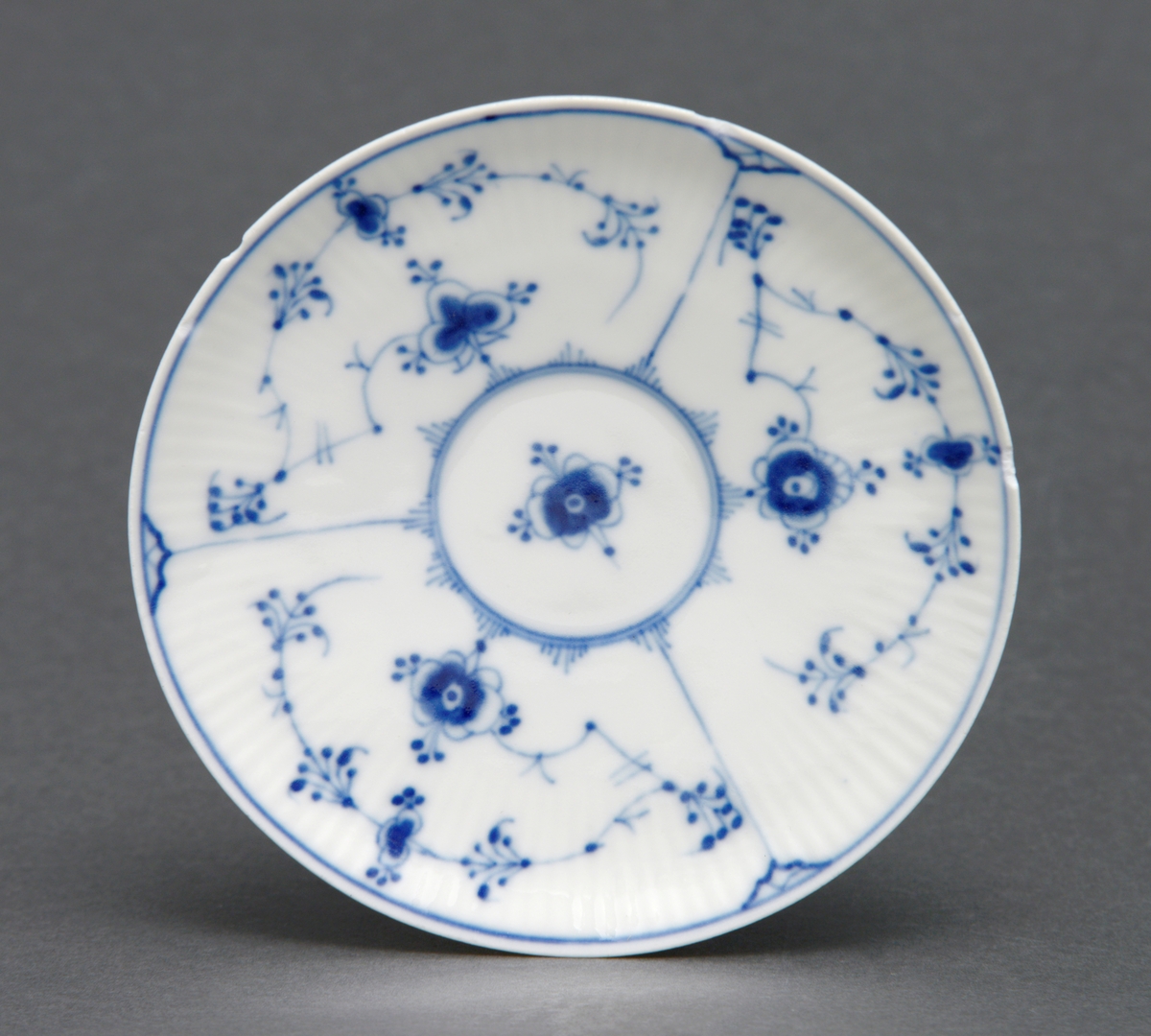 Sirkulært tefat i porselen med glasur. Rifler i porselenet og dekorert med stråmønster i blått. Tilhørende kopp (se relaterte objekter).