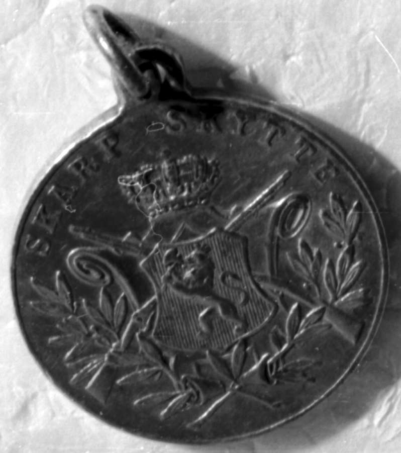 Rund medalje med det norske riksvåpenet med krone i midten over to gevær i kryss, og laurbærblad rundt.