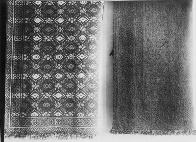 Två bohuslänska täcken (dubbelvikta). Det vänstra har mönstret som benämndes "Rosor och grenar" i spegeln. Det högra verkar vara mönstervävt med en färg på garnet så reliefverkan uppstår.