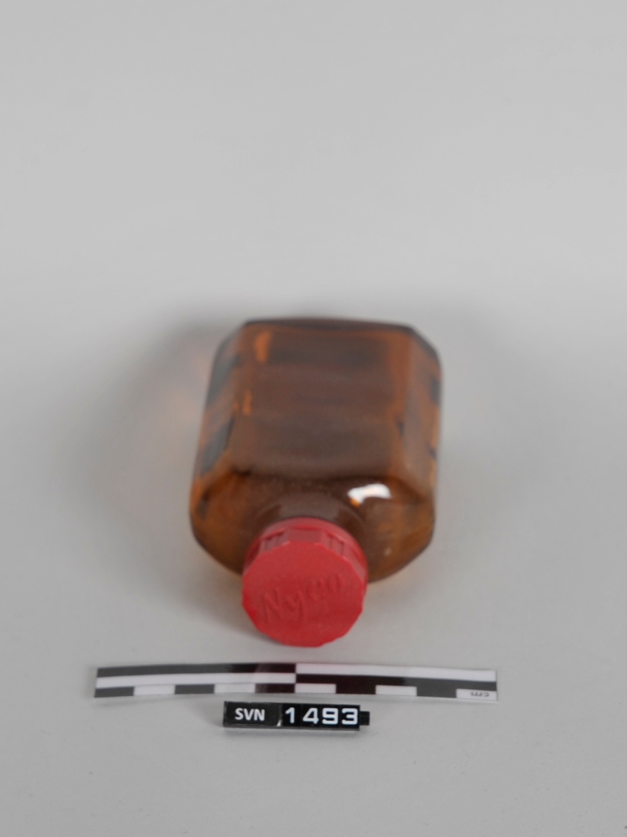 Brun oval åttekantet flaske med rød skrukork med logo. 
Flasken har ingen etikett.
På hver side av flaskens langsider er det tre(3) kortsider. Totalt to(2) langsider og seks(6) kortsider
Flaskens bunn er flat.
