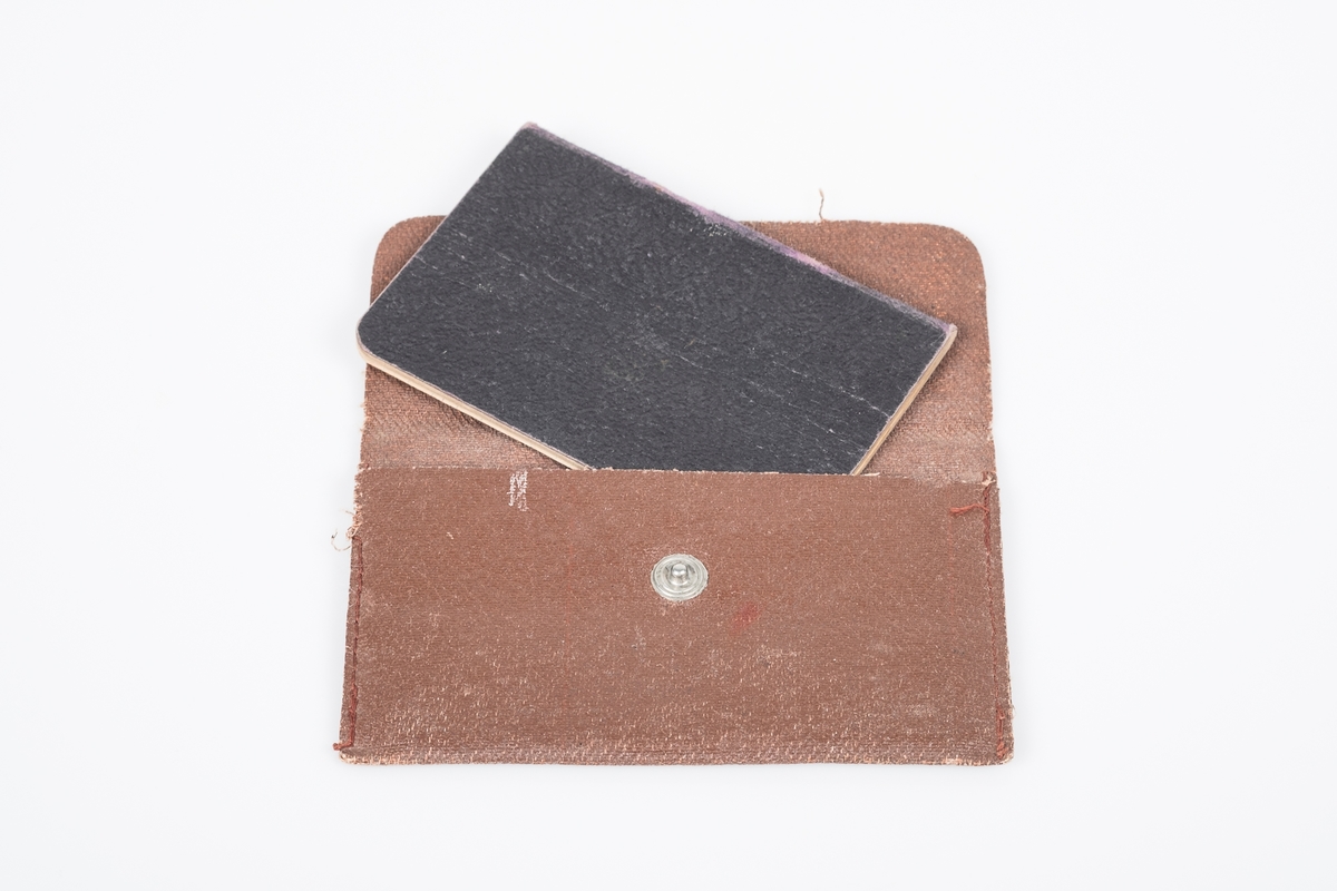 Brun flat pung i "kunstskinn", stoff med belegg og trykknapp. Inni en svart minnebok i kunstskinn, linjert papir med "autografer fra krigsfanger".