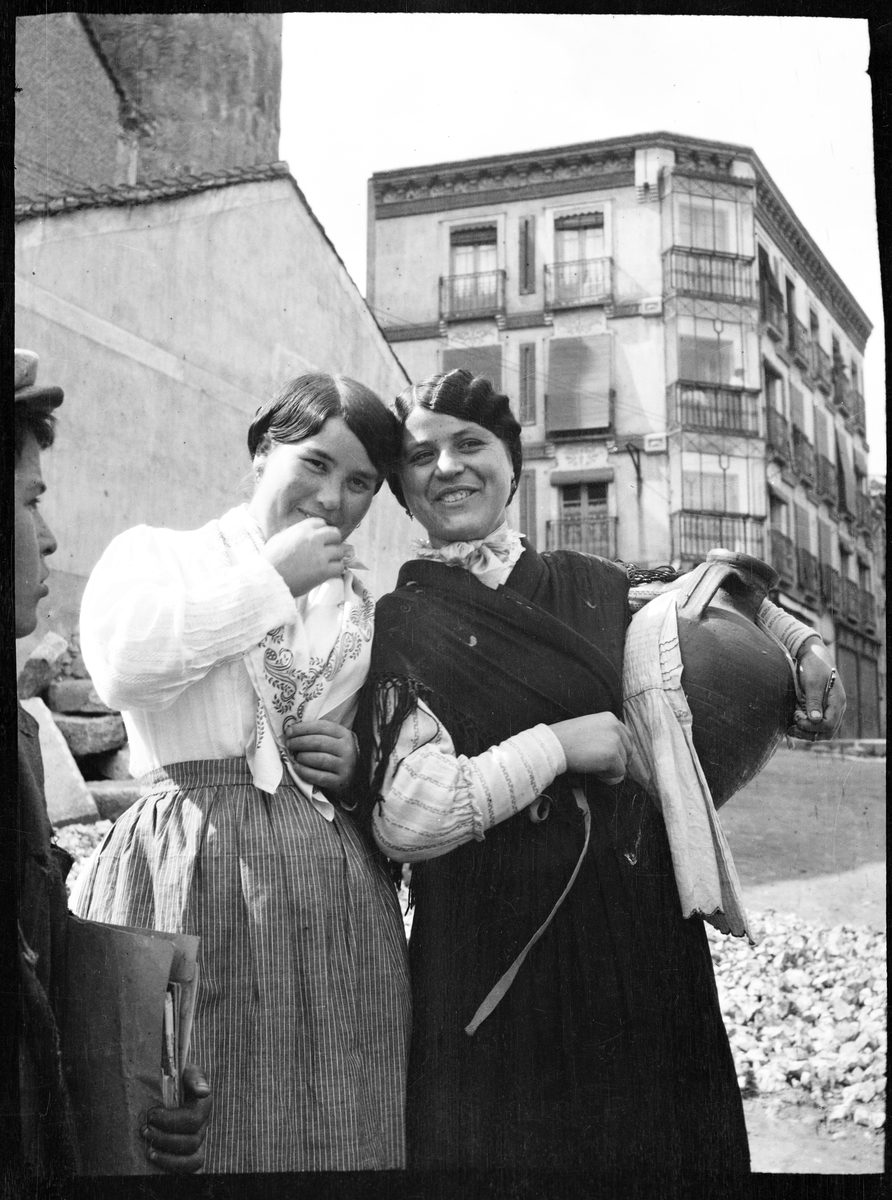 Två leende kvinnor, den ena hållande en kruka. Till vänster skymtar en pojke. Hus i bakgrunden. Troligen Ronda, Spanien.