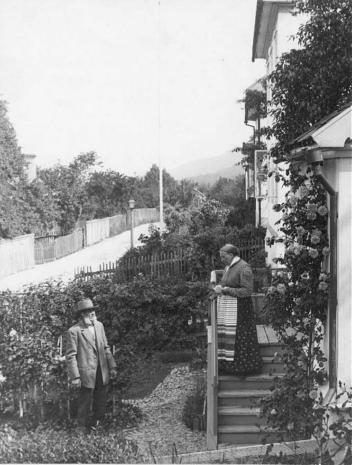 Trädgårdsmästare Olsson i trädgården vid sin fastighet på Bergsgatan 48. Hustrun Kristina Maria står på farstutrappan i prickig klänning, livstycke och randigt förkläde.