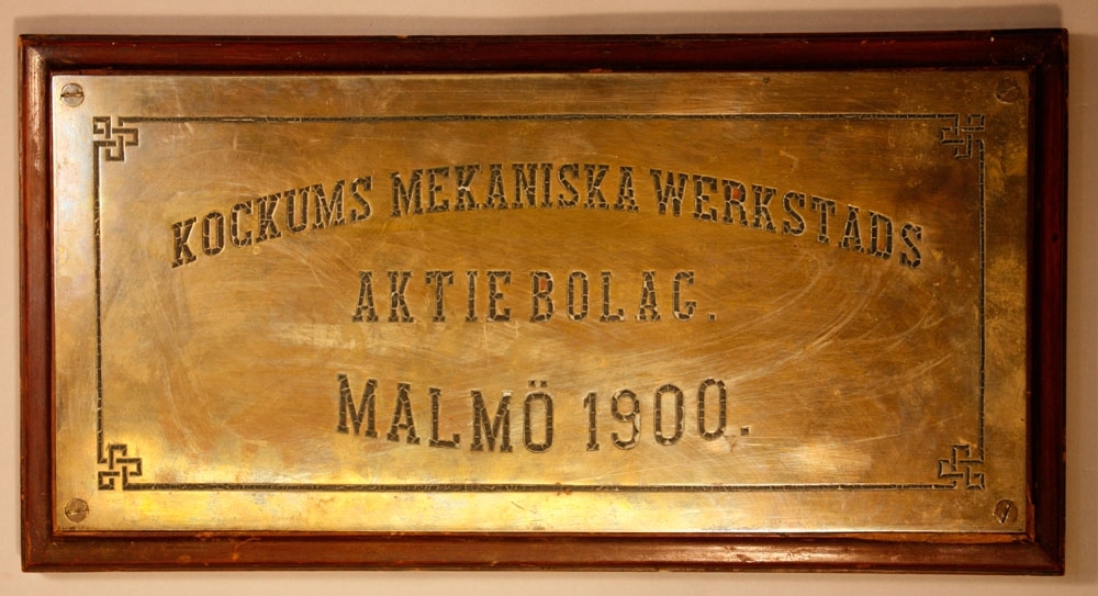 Rektangulär mässingsskylt upphängd på brun träplatta, med texten "Kockums Mekaniska Werkstads Aktiebolag Malmö 1900".
