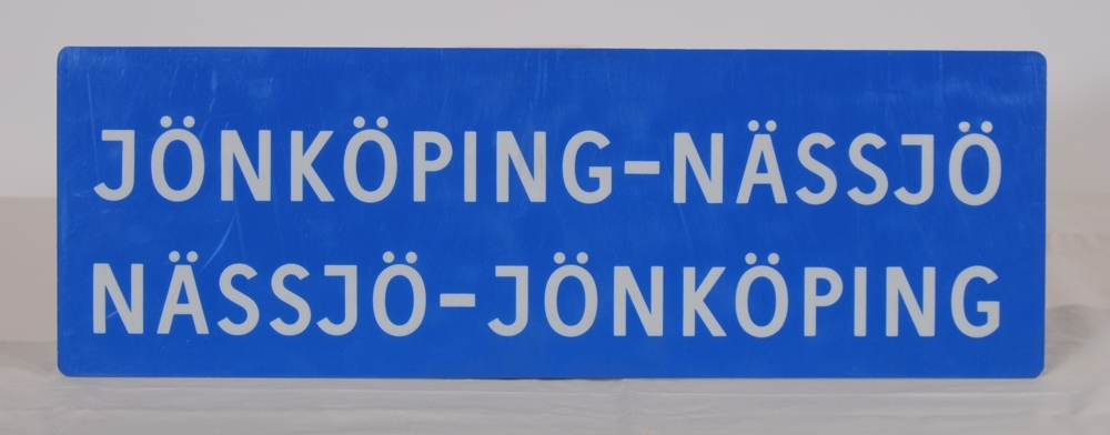 Destinationsskylt av ljusblå plast med texten "JÖNKÖPING-NÄSSJÖ NÄSSJÖ-JÖNKÖPING" i vitt på ena sidan. På motstående sida står det "NARVIK".