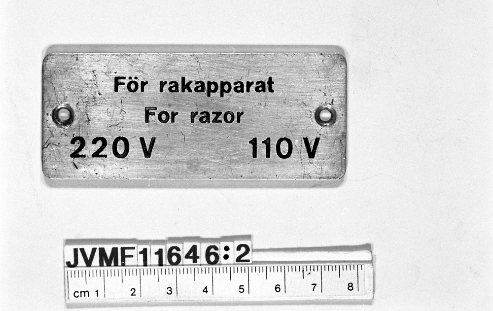 Rektangulär aluminiumsskylt med svart, försänkt text på svenska och tyska:
För rakapparat
For razor
220 V 110 V