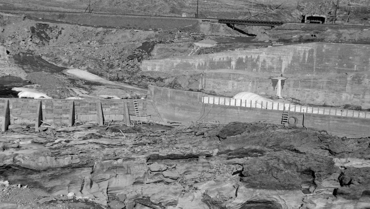 Fra dambyggingsarbeid ved Harpefoss i Gudbrandsdalslågen - i Sør-Fron kommune i Oppland fylke - vinteren 1964.  Fotografiet er tatt over et nesten tørrlagt elveleie (fra vest mot øst), mot nyoppførte murer av armert betong i et elveløp som ellers preges av opprevne bergflater.  I teksten fotografen skrev til dette bildet refereres det til en skådam, altså en ledeskjerm i elva, som var 125 meter lang og opptil 16 meter høy.  Her skulle hovedstrømmen i elva ledes mellom de to betongmurene vi ser på bildet.  I bakgrunnen (øverst i bildet) ser vi Dovrebanens trasé forbi anleggsområdet.  Kraftverket som ble bygd da dette bildet ble tatt utnytter et fall på 34 meter i Gudbrandsdalslågen.  Det ble satt i drift i 1965.  Kraftstasjonen ved Harpefossen har to kaplanturbiner og en årsproduksjon på 427 GWh.