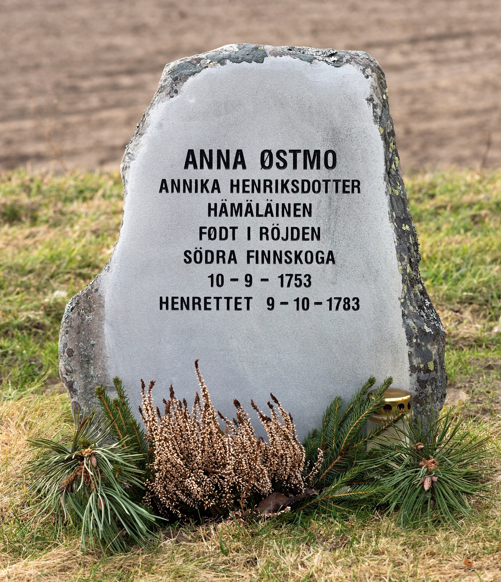 Gravstøtte, reist over Anna Østmo eller «Annika Henriksdotter Hämäläinen», som ifølge innskrift på steinen skal ha vært født i Röjden i Södra Finnskoga 10. september 1753 og død 9. oktober 1783, altså i en alder av 30 år.  Døden inntraff ved henrettelse, etter at Anna var dømt for å ha født i dølgsmål og drept barnet sitt.  Et kort resymé av saken finnes under fanen «Bildehistorikk».  I forbindelse med gravearbeid 2010 ble det funnet et hodeløst kvinneskjelett i området der man mener at henrettelsen fant sted.  Dette utløste diskusjoner om hvordan man skulle forholde seg til det man antok var levningene etter den henrettete kvinna.  Det endte med en begravelse og oppsetting av ei gravstøtte av naturstein som var slipt på fronten, som dermed fikk ei gråkvit overflate, med inngravert, svartlakkert tekst.  Da dette fotografiet ble tatt var gravstedet pyntet med ei lyngplante og litt gran- og furubar.  Vi skimter også et «gravlys», en stearinfylt sylindrisk beholder av plast med messingfarget lokk med trekkhull på sidene. 

Fotografiet er tatt som et illustrasjonsforslag til en artikkel Kari Sommerseth-Jacobsen fra Kvinnemuseet - Museene i Glåmdal har skrevet om det moderne etterspillet saken mot tjenestejenta Anna Østmo (i noen sammenhenger også stavet Anna Austmo) fikk etter skjelettfunnet i 2010.  Artikkelen var fortsatt upublisert da dette fotografiet ble registrert.