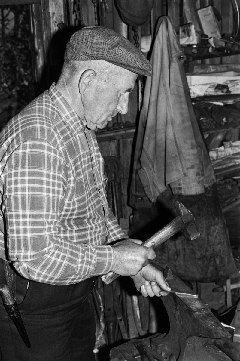 Knivsmeden Hallstein Midtbø [også kalt Hallstein Kroken] (1893-1989) fra Børtegrend i Tokke kommune i Telemark, fotografert ved smiestedet (ambolten) i smia si.  Da dette fotografiet ble tatt sto han tilsynelatende og hamret på et kaldt knivblad.  Smeden var kledd i mørke bukser med en slirekniv i beltet, han hadde rutete flanellskjorte på overkroppen og sixpencelue på hodet.  Ei arbeidsjakke hang på veggen i bakgrunn.  Vi skimter også vegghyller med ymse materiell på. 