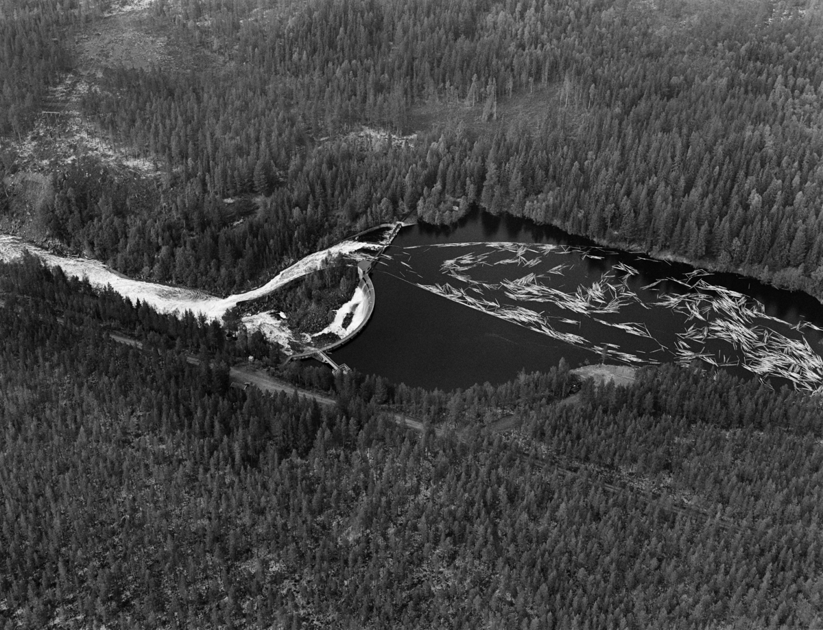 Flyfotografi tatt over kraftverksdammen i Osfallet i elva Søndre Osa i Åmot i Hedmark i mai 1984, den siste sesongen det foregikk tømmerfløting i denne delen av Glomma-vassdraget.  Fotografiet viser hvordan tømmerstokkene kom flytende med strømretningen over kraftverksdammen, der det var lagt ut lenser for å lede virket mot tømmerløpet som var plassert mot det nordre landet, hvor fallhøyden var mindre enn nedenfor den buete hoveddammen mot søndre landside.  Etter å ha passert dammen ble stokkene ført med strømmen i kvitskummende vann ned mot utløpet i elva Søndre Rena, cirka to kilometer lengre vest. Damanlegget på fotografiet skal ha vært ferdigstilt i 1947.  Den ble noe ombygd i 1981.  Ettersom vassdragslovgivinga påla dem som drev tømmerfløting å fjerne installasjoner i vann ved opphør av virksomheten, ble tømmerløpet ved Osfallsdammen revet umiddelbart etter at fløtinga på dette fotografiet var avviklet.  Energiproduksjonen ved denne lokaliteten foregår i en kraftstasjonsbygning fra 1914, cirka en kilometer nedenfor den avbildete dammen, hvor det er installert to francisturbiner som kan yte 5.5 megawatt.  Den opprinnelige dammen ble ødelagt ved et dambrudd i 1916.  Det nåværende kraftverket utnytter et fall på 41 meter i elva Søndre Osa.  Vegen på sørsida av vassdraget fører ned til kraftstasjonen og derfra mot Fylkesveg 215 ved Oset.