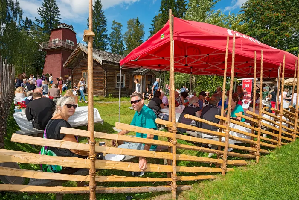 Fra friluftserveringsområdet ved skogvokterboligen fra Galten i Engerdal, der publikum tilbys vafler og kaffe under De nordiske jakt- og fiskedager på Norsk Skogmuseum i Elverum.  Området er inngjerdet med skigard der det er brukt gransveig som bindsel.  Publikum sitter ved bord og benker langs skigarden, enkelte under et rødt tekstilt tak fra Coca Cola.  I bakgrunnen skimtes den nevnte skogvokterboligen og et brannvakttårn som er rekonstruert med et originaltårn fra Haukenesfjellet i Rømskog i Østfold som forbilde.  Fotografiet ble tatt søndag 10. august 2014.