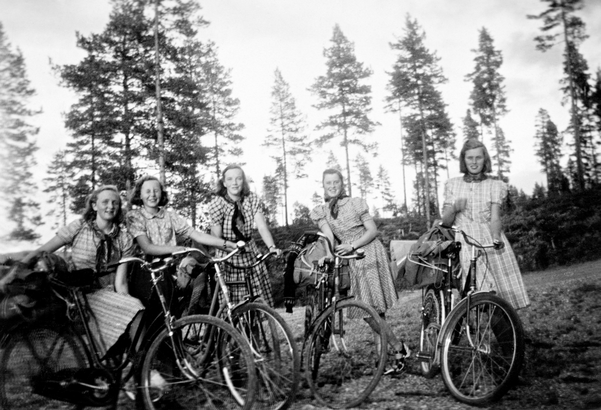 Friluftsliv under 2. verdenskrig.  Fotografiet viser fem elverumsjenter som syklet til Rysjøen i Trysil (Søre Osen) for å nye samværet der noen dager.  Jentene poserer ved en vegkant med hver sin sykkel, og med ryggsekker som er festet til bagasjebrettene ved hjelp av reimer.