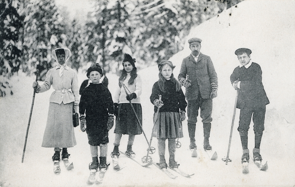 Postkort. Motivet på kortets fremside viser en mann, en kvinne og fire barn på skitur i vinterlandskap. Skigåing.