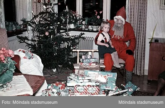 Jultomte med en flicka i knät och julklappar framför sig. Foto 1 är sv/v och foto 2 är i färg. Okända personer, plats och årtal.