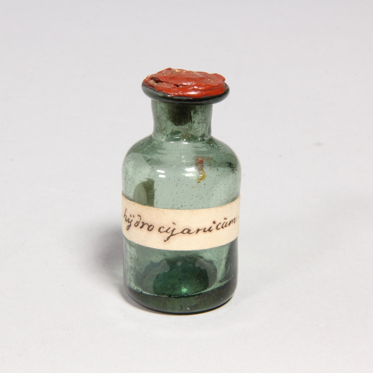 Flaskor, 10 st, av grönt glas med proppar av kork förseglade med rött lack. Står i pappask med lock klädd med marmorerat papper. Flaskorna innehåller rester av cyanid.