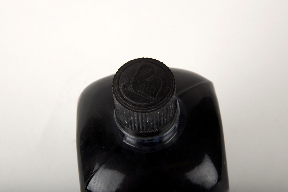 Glassflaske fylt med svart skriveblekk. Flasken har kork av plast med indenterte linjer. På toppen av korken er et uthevet bilde av en fugl som står over to fugleunger. Hvit etikett med blå tekst og ramme på fremsiden.