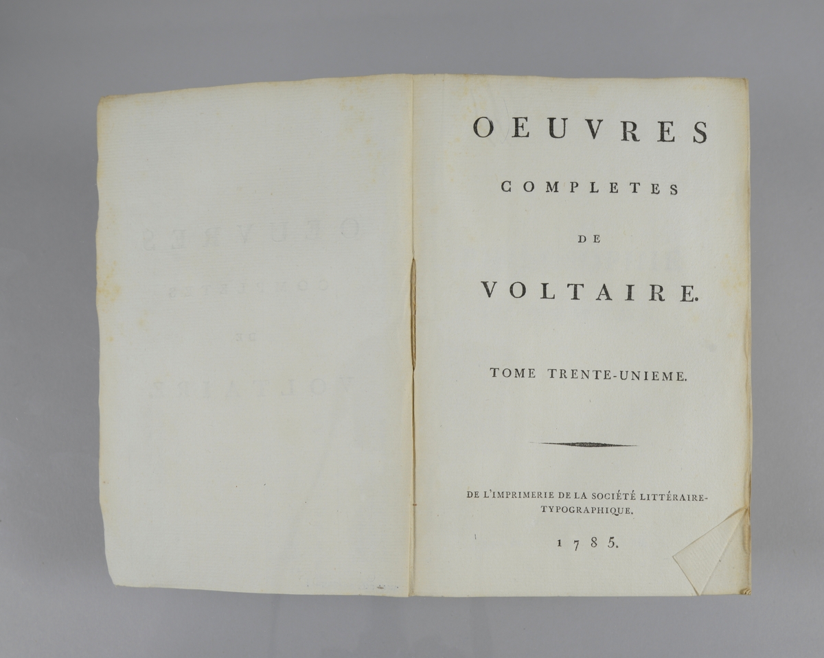 Bok, pappband,"Oeuvres completes de Voltaire", del 31 "Philosophie de Newton", tryckt 1785.
Pärmen klädd med gråblått papper, på pärmarnas insidor klistrade sior ur annan bok. Med skurna snitt. På ryggen klistrad pappersetikett med tryckt text samt volymens nummer. Ryggen blekt. Med planscher.