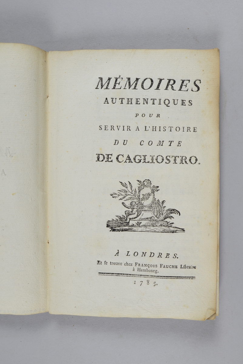 Bok, pappband, "Mémoires authentiques pour servir à l´histoire du comte de Cagliostro", tryckt 1785 i London. Pärmar av gråblått papper, på insidan klistrade sidor ur annan bok. På ryggen bokens titel samt etikett med samlingsnummer. Skuret snitt.