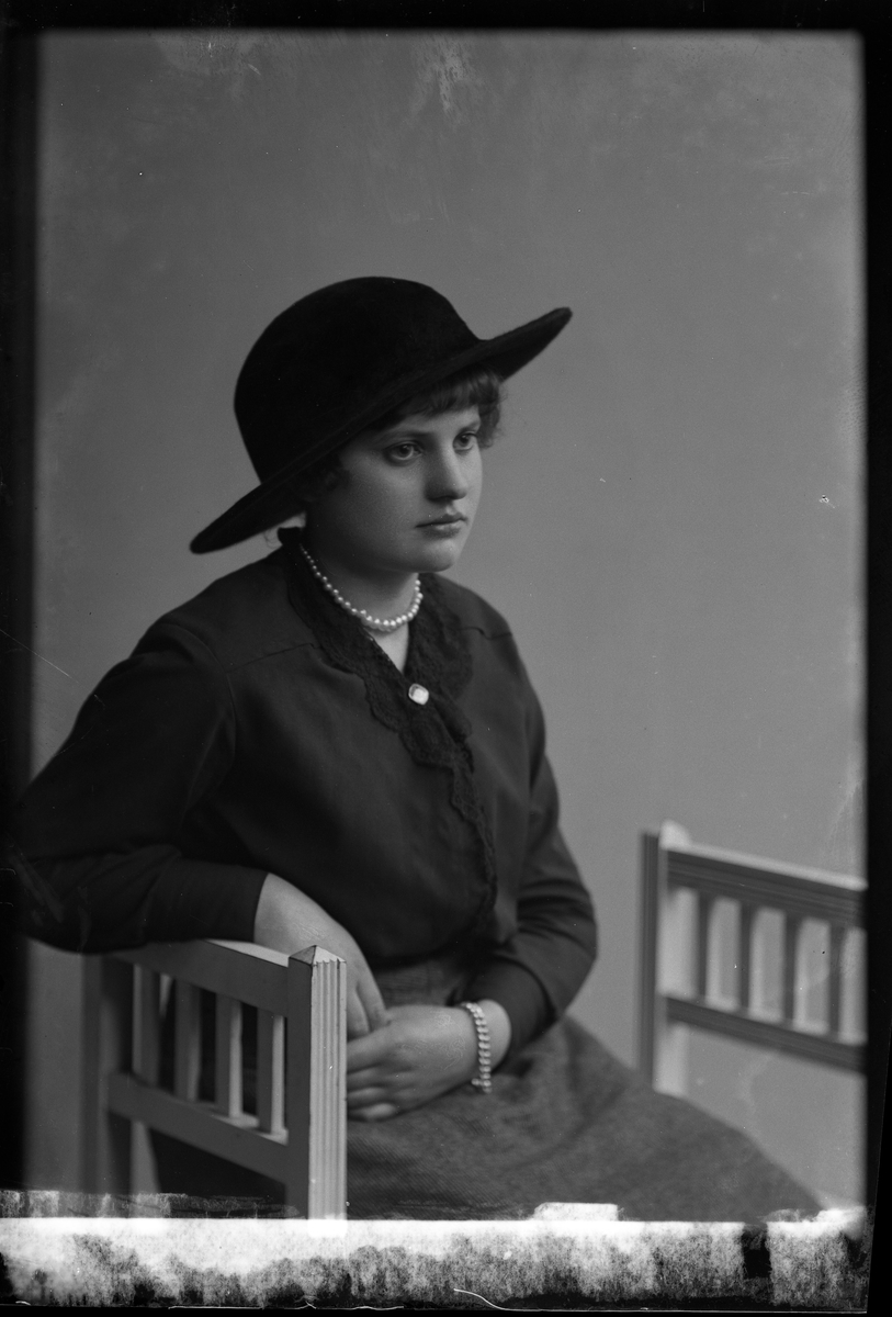 Porträtt av Vivan Hedlund sittandes på en pall och klädd i svart bredbrättad hatt samt mörk blus och kjol. Runt handled och hals bär hon pärlor.