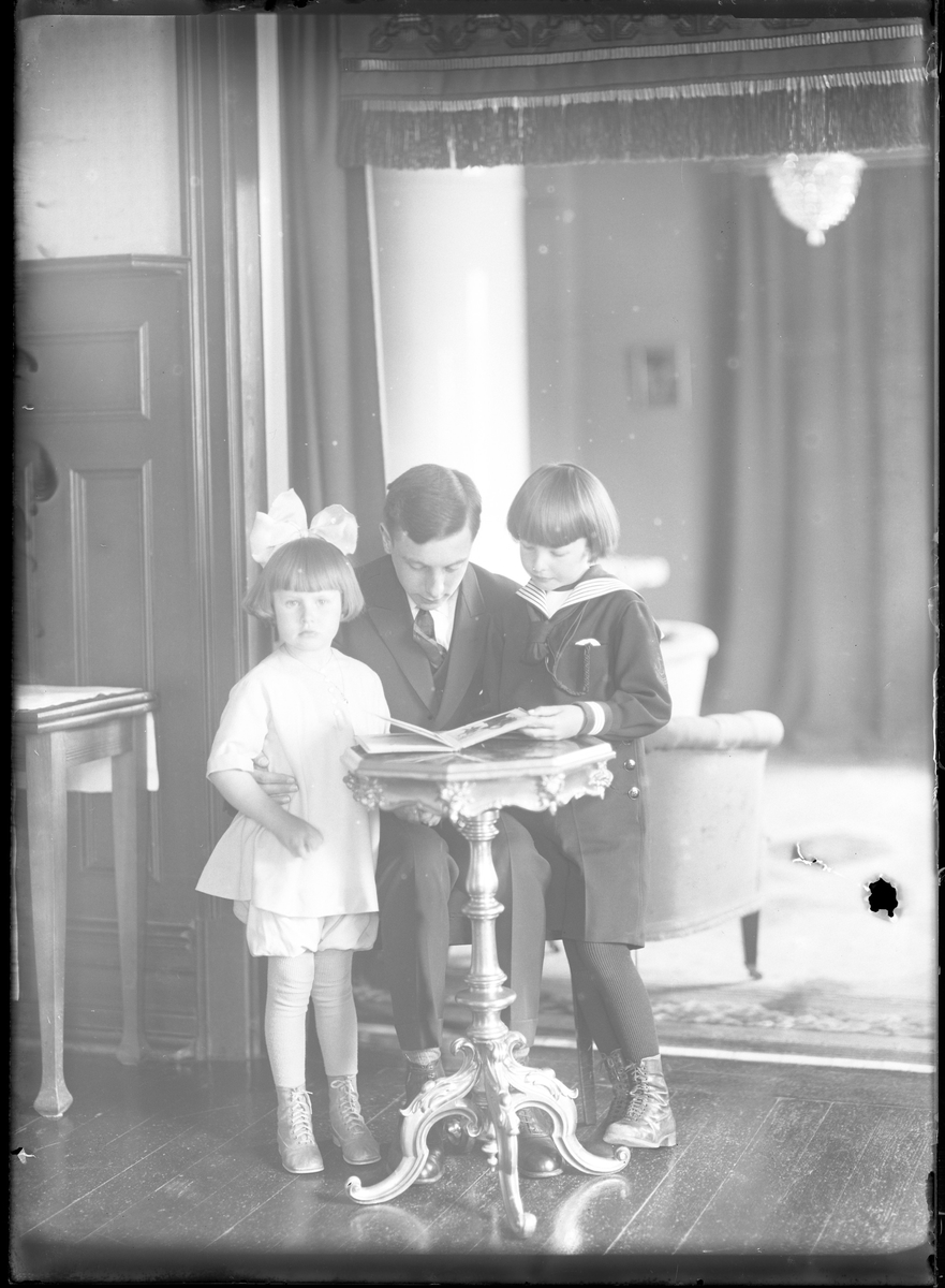 En kostymklädd ung man sitter på en stol och bredvid honom står en pojke i sjömanskläder och en flicka i ljus klänning och rosett i håret. Tillsammans tittar de i en bok. I fotografens egna katalog står det "Barnen Norinder".