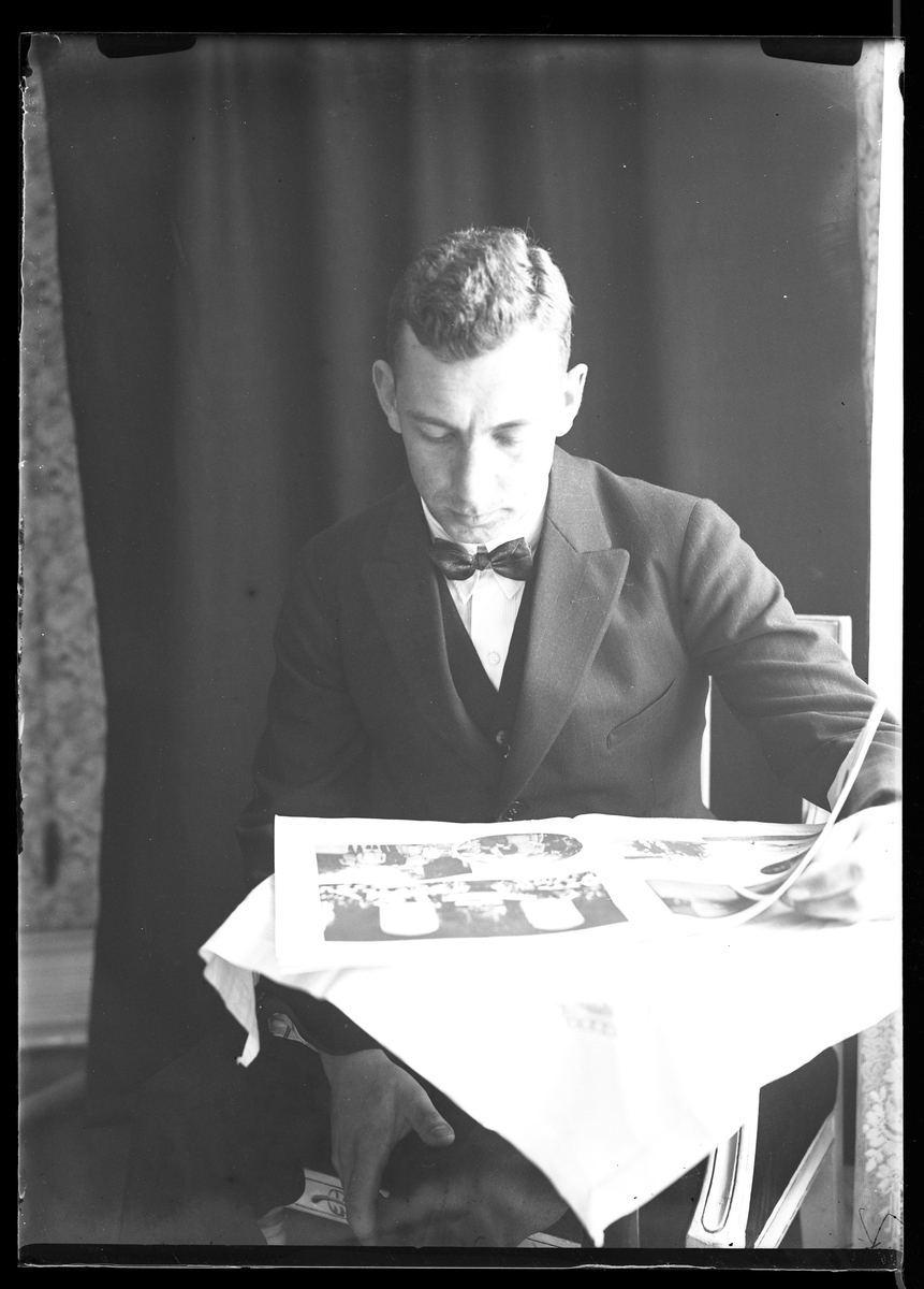 Porträtt av fotografen själv när han sitter vid ett bord och läser tidningen. Bakom honom hänger ett svart draperi.