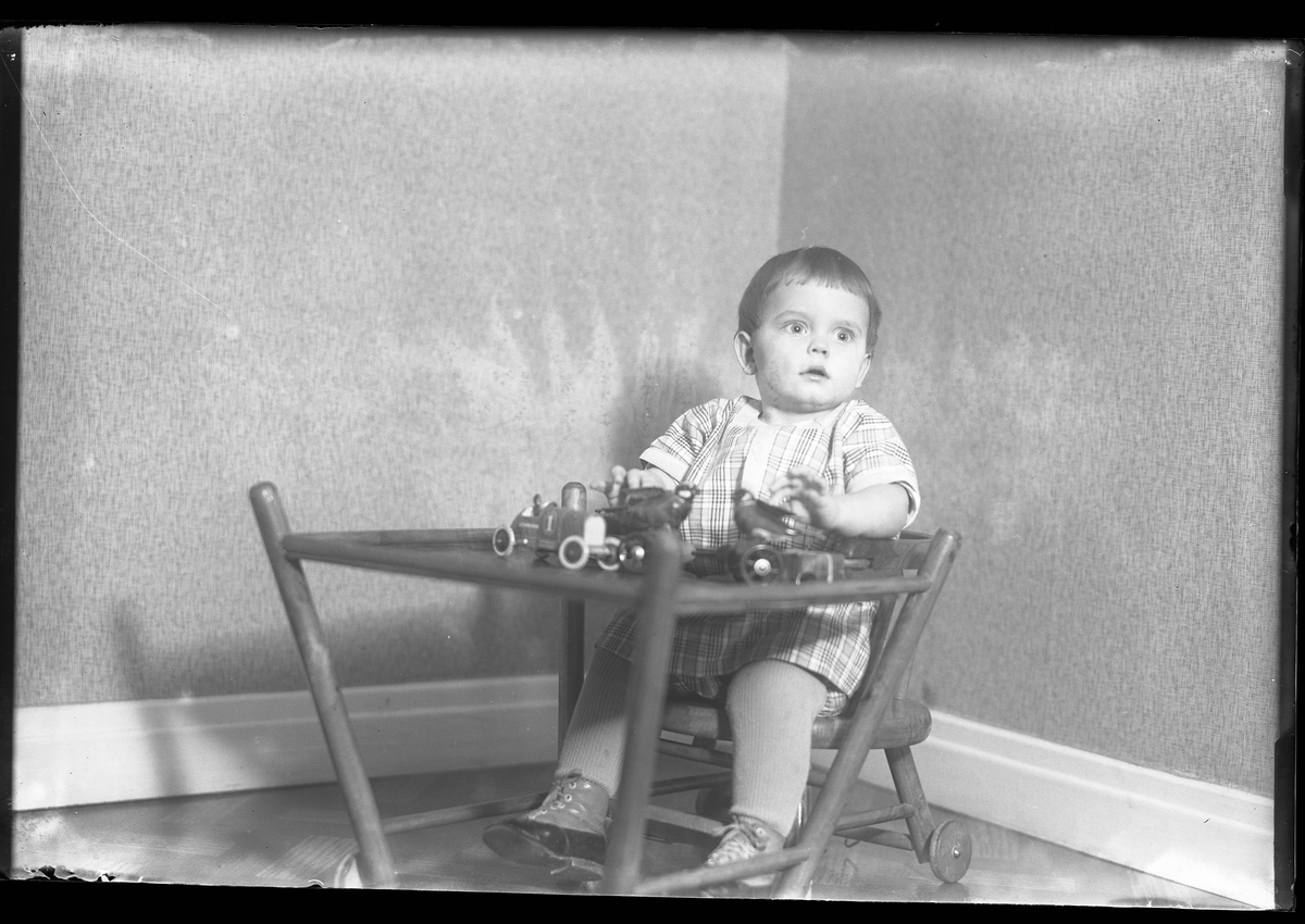 En liten pojke i rutig klänning, stickade strumpor och kängor sitter i en nedfälld barnstol och leker med plåtleksaker. I fotografens egna anteckningar står det "Gust. Claessons pojke"