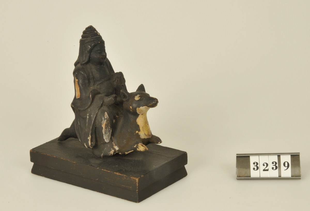 Statyett skuren i trä. 
Buddhafigur ridande på en hund. 
Målad i svart.

Har tillhört de Adelskölska samlingarna.
