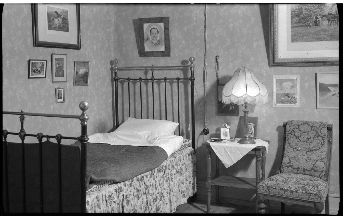 Interiör från en äldre bostadshus, villa Charlottenberg (Hedvigsberg) vid Göteborgsvägen. Huset byggt på 1800-talet.
Sovrum med järnsäng som står i ett hörn.
Flera tavlor hänger på väggen över sängen.

Sängen har blommig sängkappa och är bäddad med kudde, lakan och filt eller täcke.
Ett nattduksbord med bordslampa med fransar och ett par fotografier.
Till höger om nattduksborden en stoppad stol utan armstöd.
Blommönstrad tapet.


Elin Bothén.

Foto 13 september 1966.