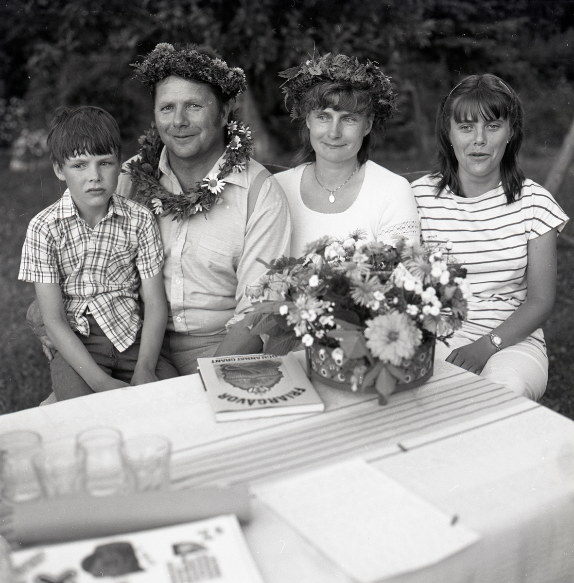 En familj sitter vid ett bord med duk, blombukett, glas och boken "Friargåvor" den 30 juli 1982. Några av dem bär blomkransar på huvudet.