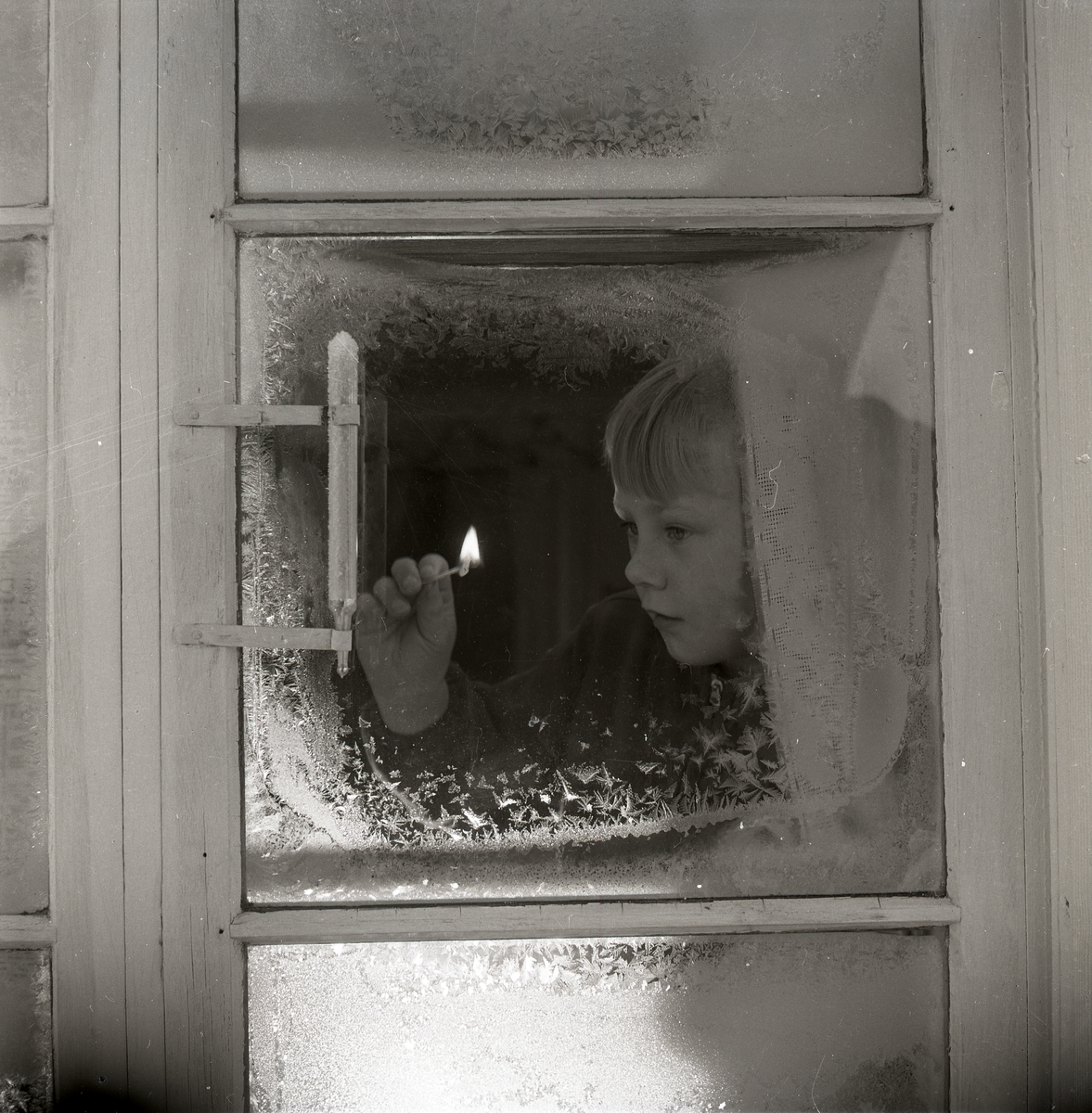 Innanför ett fönster tänder en pojke en tändsticka, för att bättre kunna se på utetermometern hur många grader det är. Fönstret är täckt med rimfrost, 4 januari 1953.