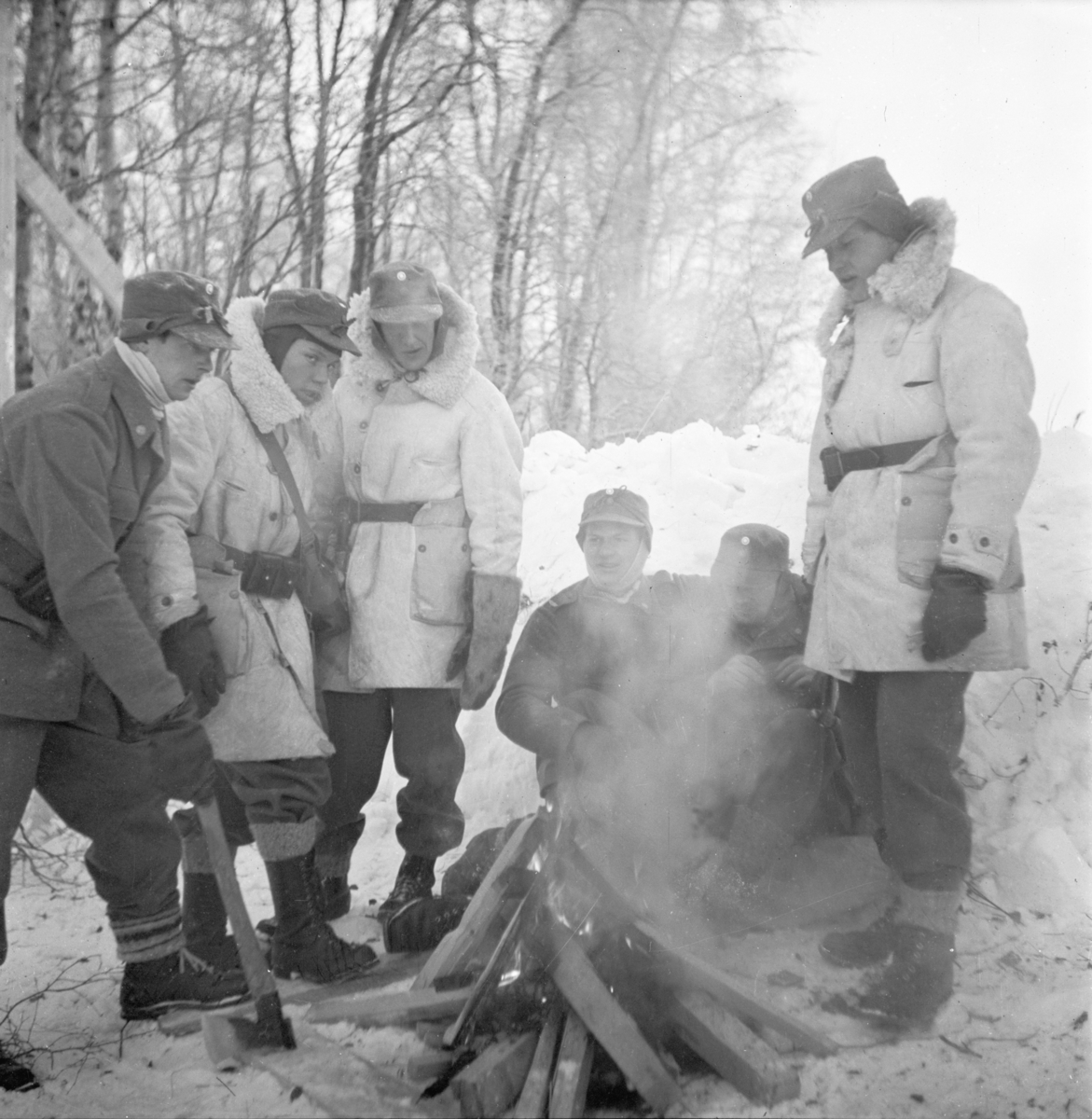 Militärer värmer sig vid brasa vid Svenska frivilligkåren i Finland, F 19. Sex militärer i uniform vid öppen eld i terräng.