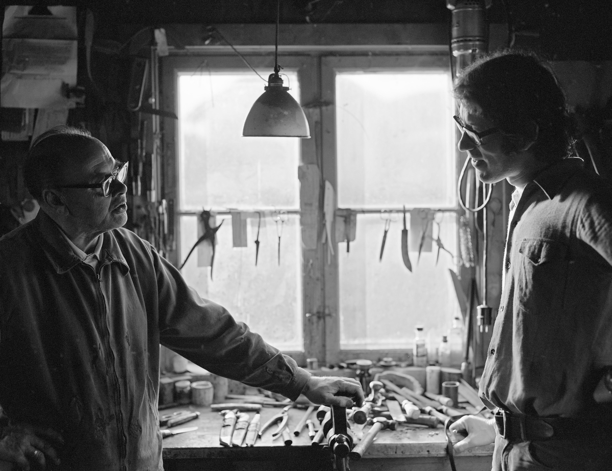 Knivmakeren Even Johannes Blindingsvolden (1908-1985) fra Rudshøgda i Ringsaker, til venstre, i samtale med daværende vitenskapelig assistent ved Norsk Skogbruksmuseum, Åsmund Eknæs (1940-1981).  Fotografiet ble tatt høsten 1973, da Eknæs besøkte Blindingsvolden for å dokumentere hvordan han arbeidet med knivproduksjonen i verkstedet på småbruket Rudsland.  I bakgrunnen ser vi en sentral del av arbeidsbenken til knivmakeren.

Mer informasjon om knivsmeden Even Blindingsvolden og museumsmannen Åsmund Eknæs finnes under fanene «Bildehistorikk» og «Opplysninger».